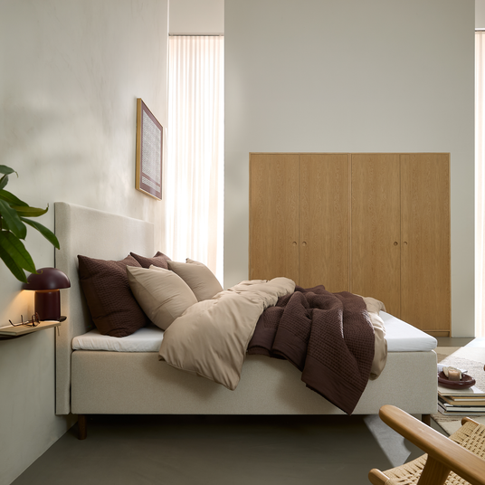 Soveværelse med beige seng, brune og beige dyner og puder, lyse træskabe og blødt dagslys gennem skærmede gardiner; bordlampe og plante ved siden af.