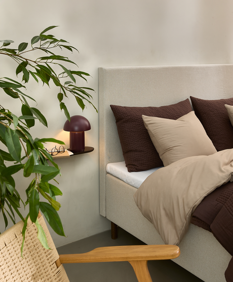Roligt soveværelseshjørne med beige seng, brune og beige puder, bordeaux bordlampe på lille væghylde, træstol og grøn plante.