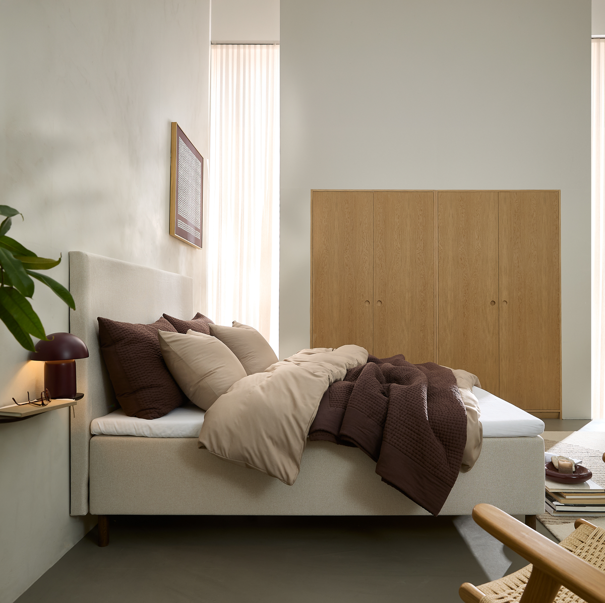Beige polstret seng med brunt og beige sengetøj i et roligt soveværelse, blødt dagslys fra høje gardiner, indbyggede garderobeskabe i træ, lille bordlampe og grøn plante.
