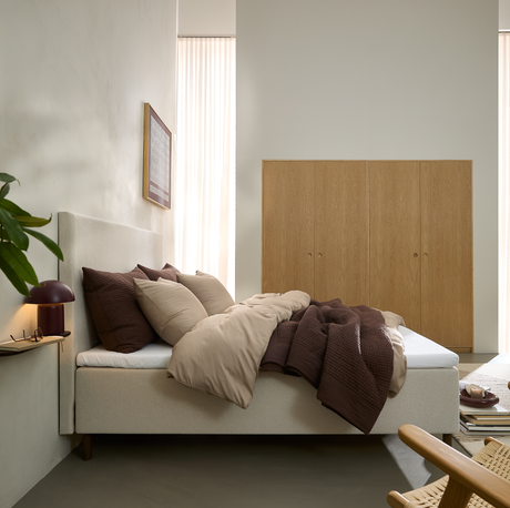 Beige polstret seng med brunt og beige sengetøj i et roligt soveværelse, blødt dagslys fra høje gardiner, indbyggede garderobeskabe i træ, lille bordlampe og grøn plante.