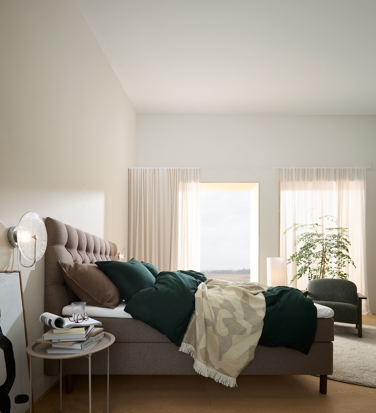 Lyst og roligt soveværelse med polstret seng i brune nuancer, grønne puder og plaid, lyse gardiner, væglampe, lille sidebord med bøger og glas, grøn plante og lænestol ved vinduet i dagslys.