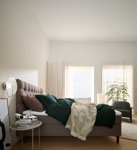 Lyst og roligt soveværelse med polstret seng i brune nuancer, grønne puder og plaid, lyse gardiner, væglampe, lille sidebord med bøger og glas, grøn plante og lænestol ved vinduet i dagslys.