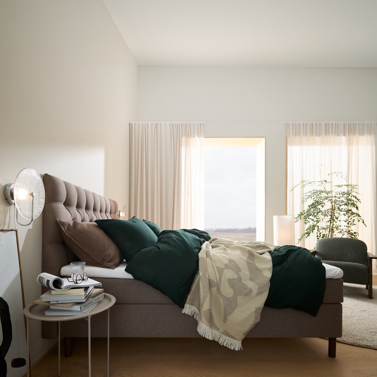 Lyst soveværelse med polstret seng, mørkegrønt sengetøj og beige plaid, lyse gardiner, natbord med bøger og glas samt en grøn plante ved vinduet.