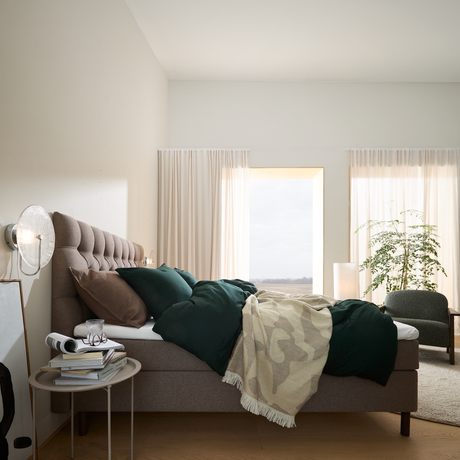Lyst soveværelse med polstret seng, mørkegrønt sengetøj og beige plaid, lyse gardiner, natbord med bøger og glas samt en grøn plante ved vinduet.