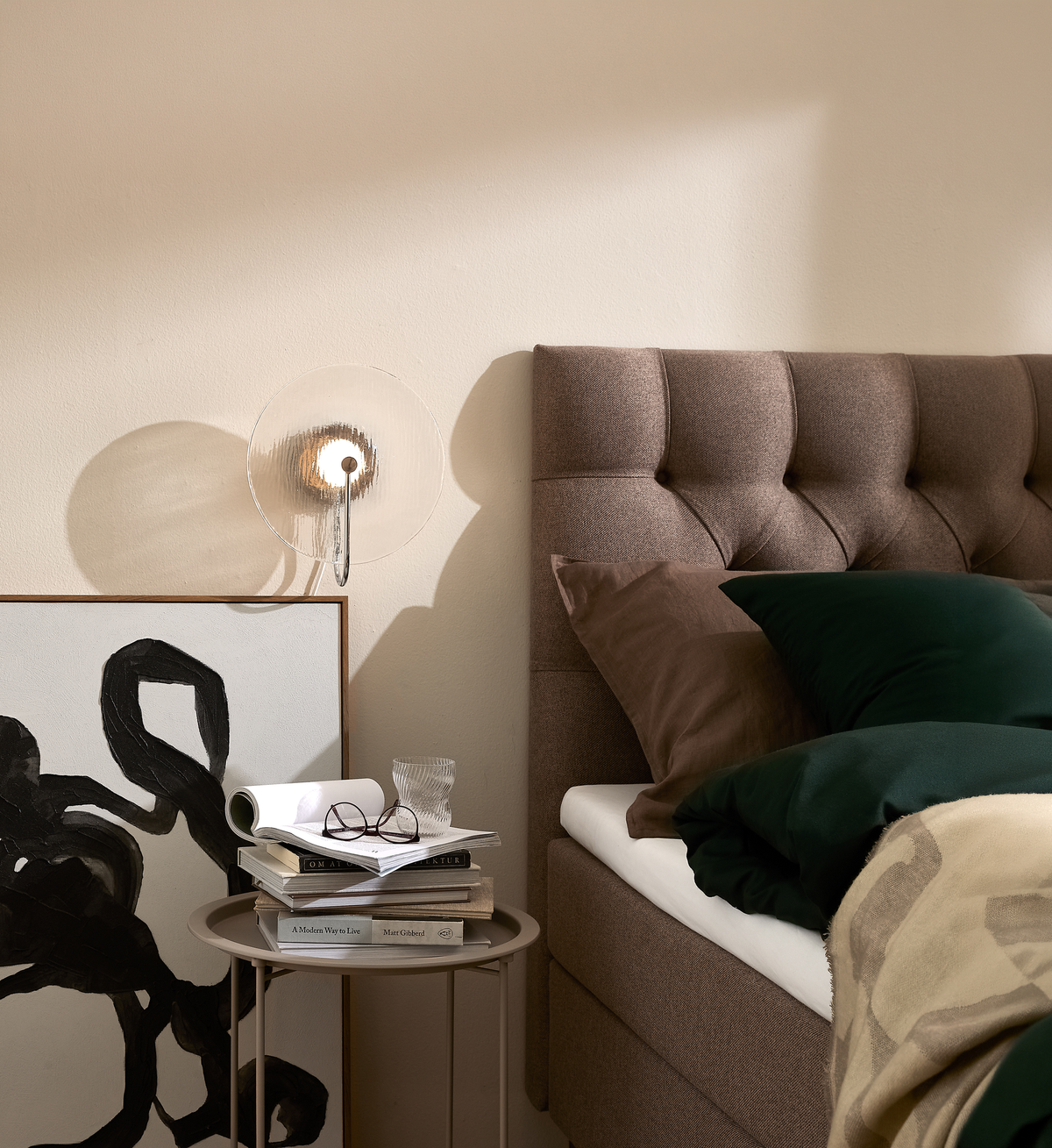 Soveværelse med brun polstret sengegavl, grønne og brune puder, beige plaid, rundt sidebord med glas, briller og bøger inklusiv “A Modern Way to Live”, abstrakt sort‑hvid kunst og glas væglampe i blødt dagslys.