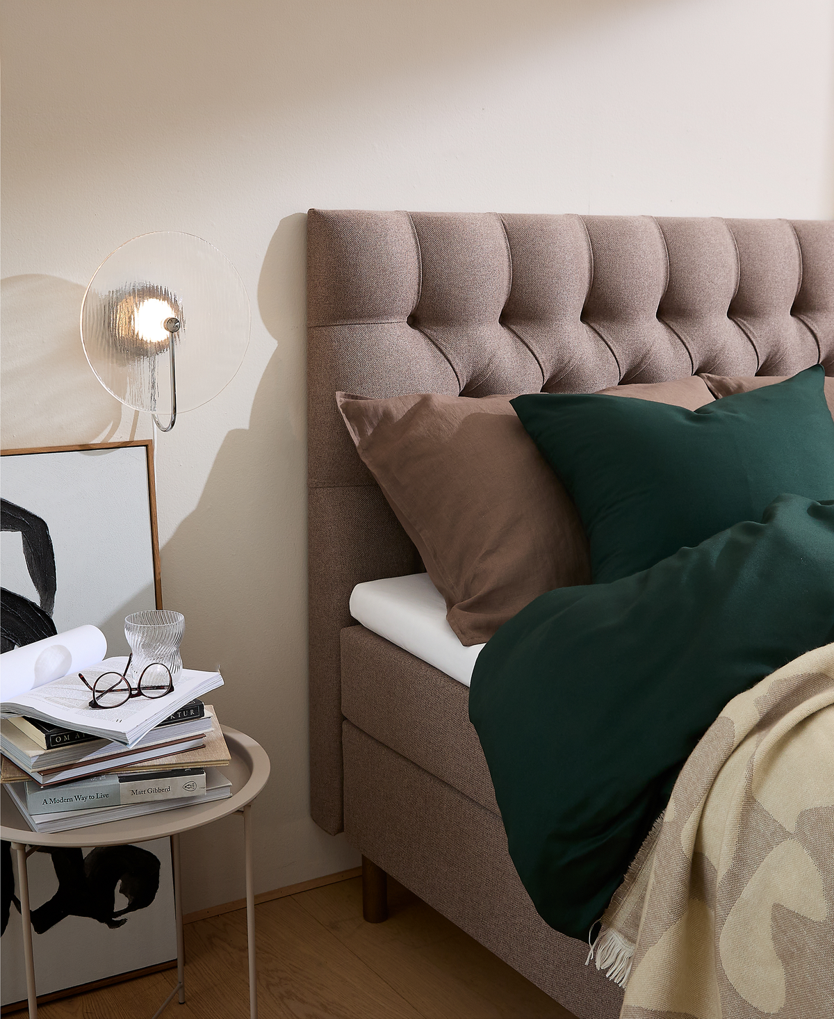 Soveværelse med polstret, knapstoppet sengegavl i beige, grønt dynebetræk, brune puder og plaid; vægmonteret glaslampe lyser over et lille sidebord med glas, briller og bøger, bl.a. A Modern Way to Live, samt indrammet sort‑hvid kunst.