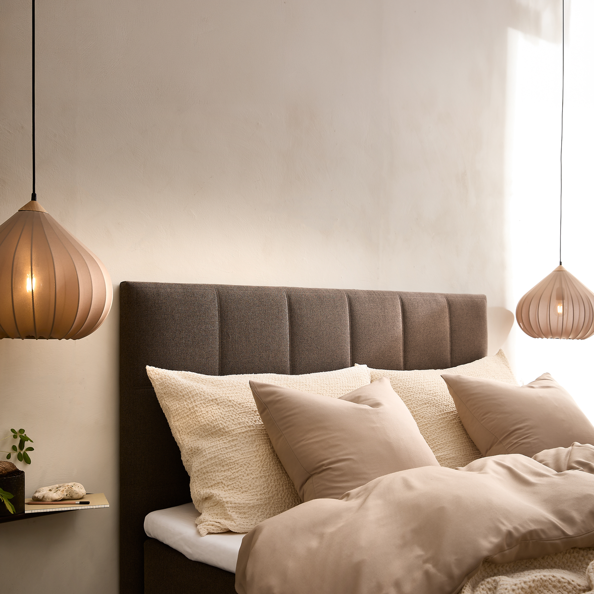 Hyggeligt soveværelsesmiljø med brunpolstret sengegavl, beige sengetøj og to beige pendellamper, blødt dagslys på en neutral væg samt en lille hylde med plante og bog.