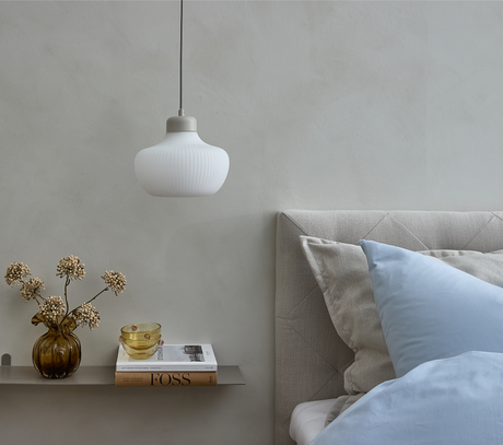 Rolig soveværelsesscene med beige sengegavl, lyseblåt sengetøj, hvid pendellampe, ravfarvet vase med tørrede blomster, to skåle og bogen FOSS på en smal væghylde.