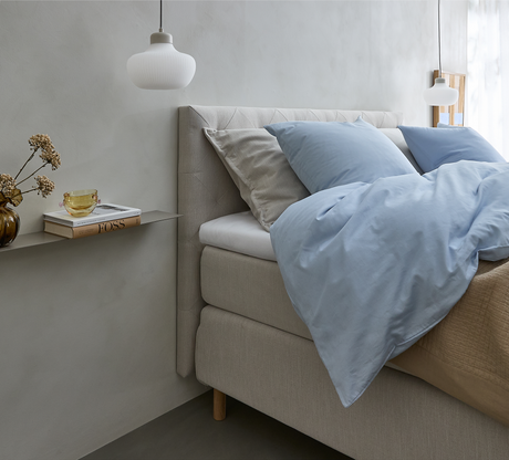 Lyst soveværelse med beige seng, lyseblåt sengetøj og puder, to hvide pendler samt en smal hylde med vase, gul kop og bogen FOSS ved siden af sengen.