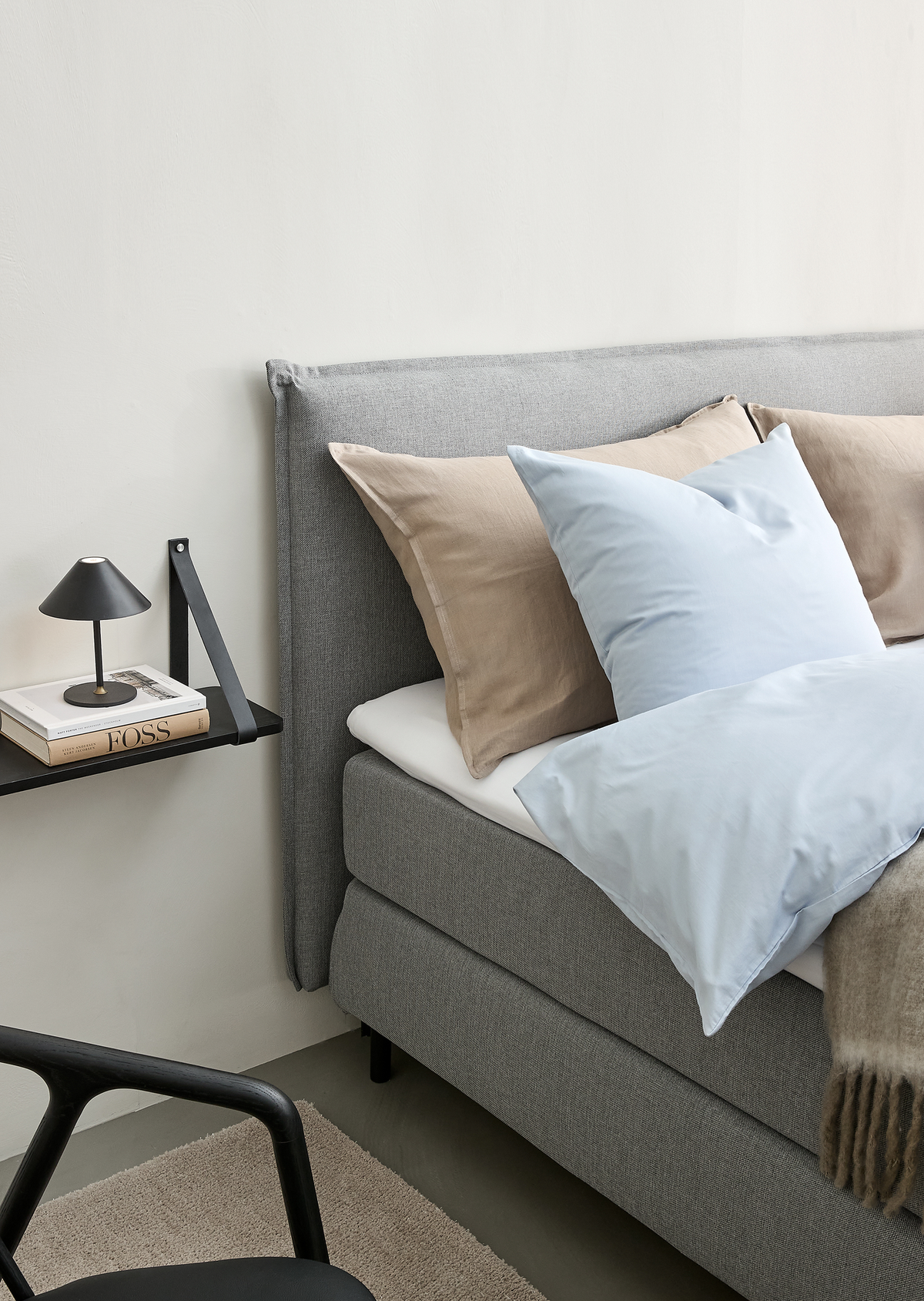 Grå polstret seng med beige og lyseblå puder og dyne; på væghylden ved siden af står en lille sort lampe og bøger, bl.a. FOSS.