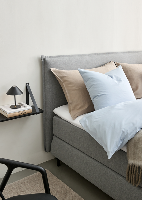 Grå polstret seng med beige og lyseblå puder og dyne; på væghylden ved siden af står en lille sort lampe og bøger, bl.a. FOSS.