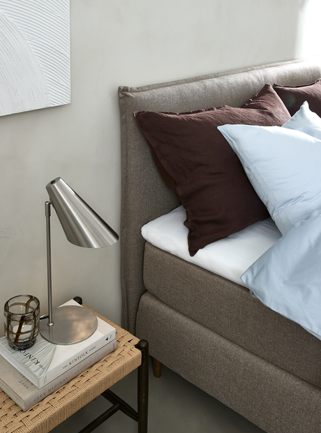 Nærbillede af et senges hjørne med grå polstret gavl, brune og lyseblå puder samt et sengebord med stålbordlampe, glasfyrfadsstage og bøgerne The Kinfolk Home og Soft Minimal.