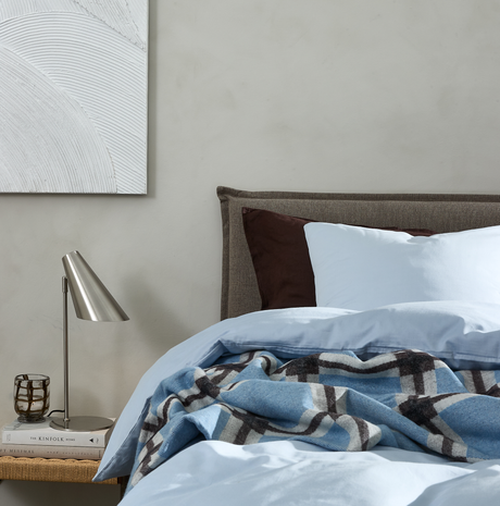 Soveværelsesscene med lyseblåt sengetøj, rutet blå plaid over sengen, brun pude og polstret grå sengegavl, samt natbord med metallisk bordlampe, glas og bogen The Kinfolk Home.