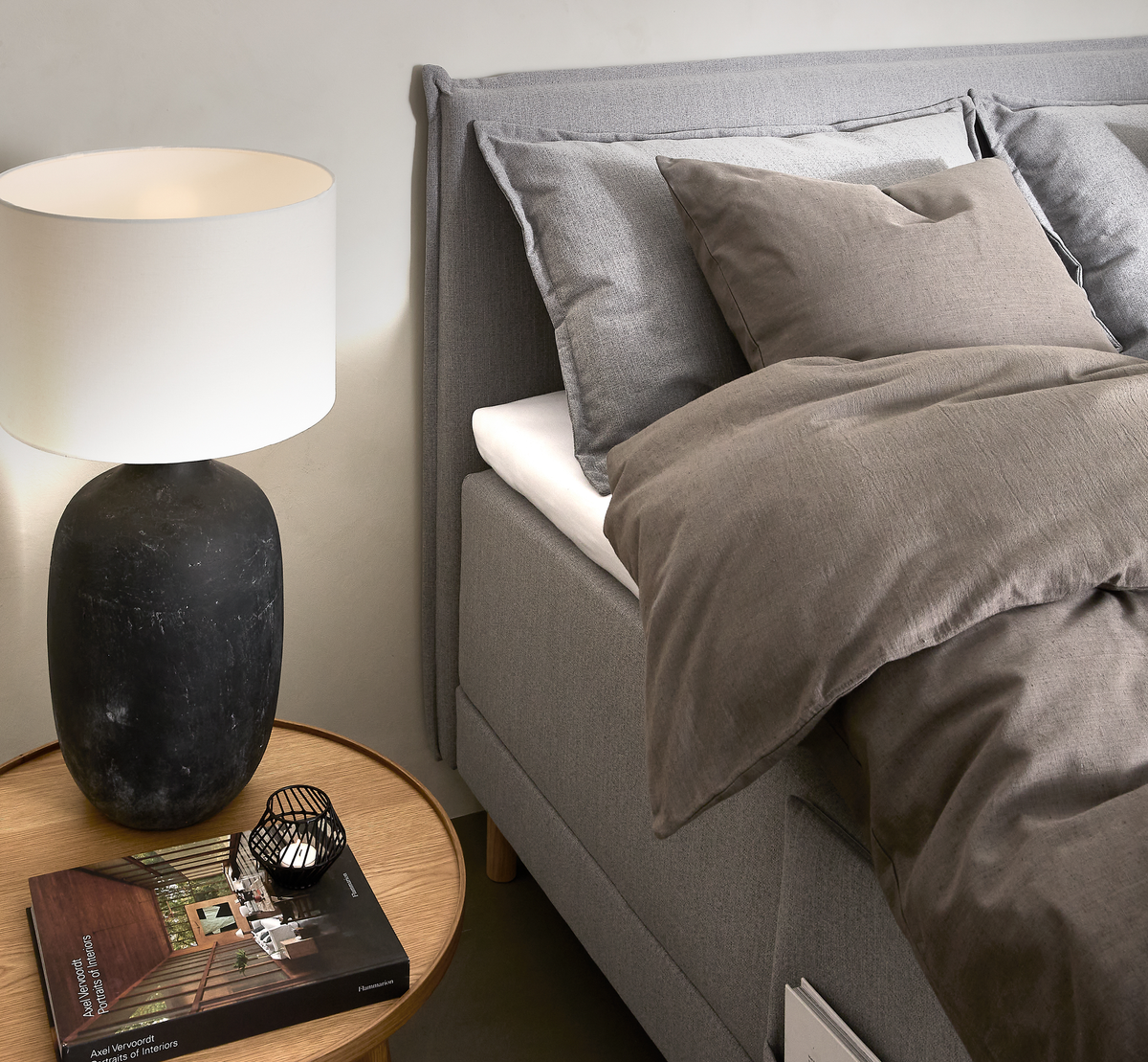 Soveværelse med grå seng, beige‑brunt sengetøj, rundt egetræsnatbord med sort keramiklampe, lille fyrfadsstage og bogen Axel Vervoordt: Stories of Interiors.