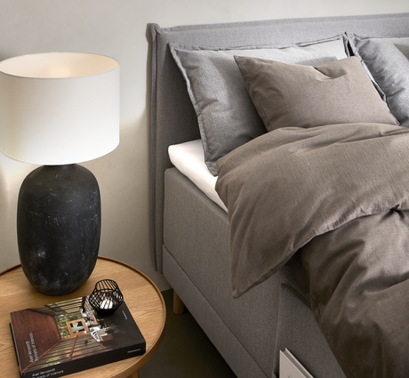 Soveværelse med grå seng, beige‑brunt sengetøj, rundt egetræsnatbord med sort keramiklampe, lille fyrfadsstage og bogen Axel Vervoordt: Stories of Interiors.