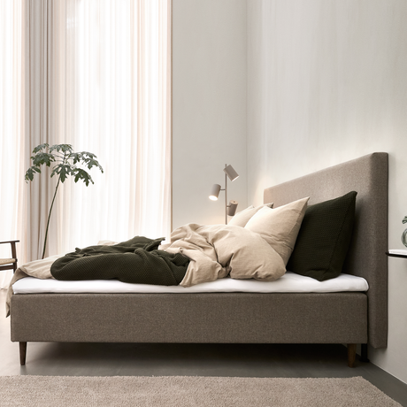 Minimalistisk soveværelse med beige polstret seng, hørfarvede puder, mørkegrønt plaid, gulvlampe og lette gardiner med dagslys.