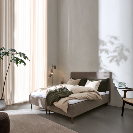 Lyst soveværelse med polstret seng i beige og grønne nuancer, lette gardiner, væglampe, sidebord med glasvase og en stor plante i hjørnet.