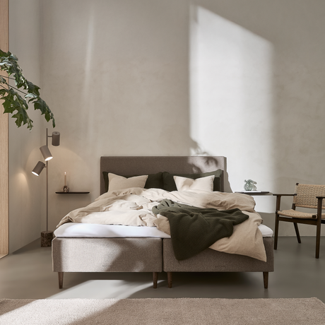 Lyst soveværelse med sollys på væggen, grå polstret seng med beige sengetøj og grøn plaid, plante til venstre, lille sidebord med lys og stol i flet.