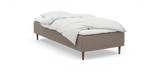 En enkeltseng med beige stofbase, træben og hvidt sengetøj; diskret nocturne‑logo på siden._ps