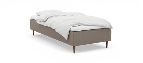 En enkeltseng med beige stofbase, træben og hvidt sengetøj; diskret nocturne‑logo på siden.