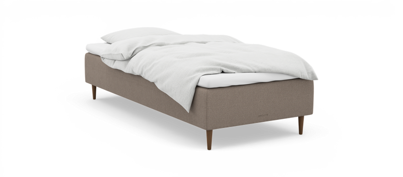 En enkeltseng med beige stofbase, træben og hvidt sengetøj; diskret nocturne‑logo på siden._ps