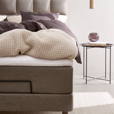 Moderne seng i beige tekstil med cremefarvet strikket plaid og mørklilla sengetøj; på et smalt sengebord ligger en bog med titlen FOSS og en lilla glasvase.