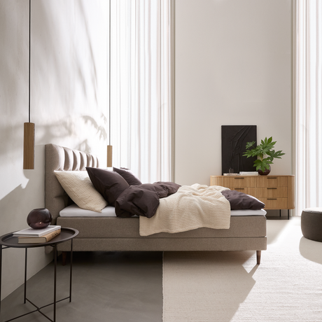 Lyst, minimalistisk soveværelse med beige polstret seng, mørkebrune puder og cremefarvet plaid; to træpendler, sort sidebord med bog og glasdekoration samt lav trækommode med grøn plante ved store, lyse gardiner i dagslys.