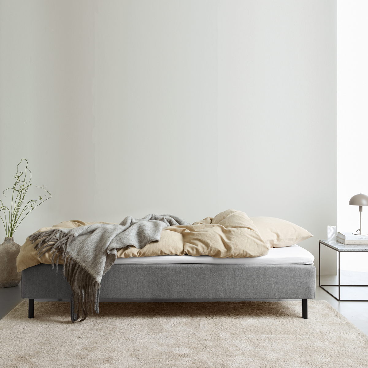 Grå polstret seng med beige sengetøj og grå plaid; natbord med lampe og bøger i et lyst, minimalistisk soveværelse.