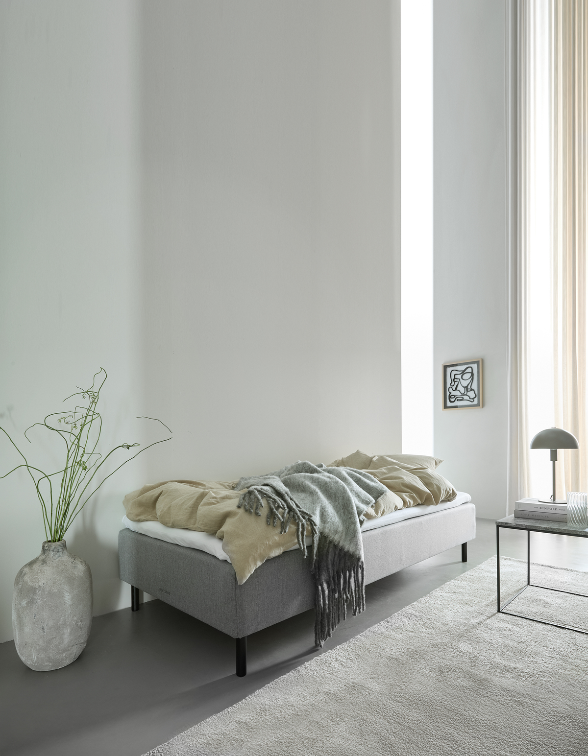 Lyst soveværelse med enkel grå seng, beige og grå plaid, plante i vase, bordlampe og blødt dagslys fra høje vinduer.