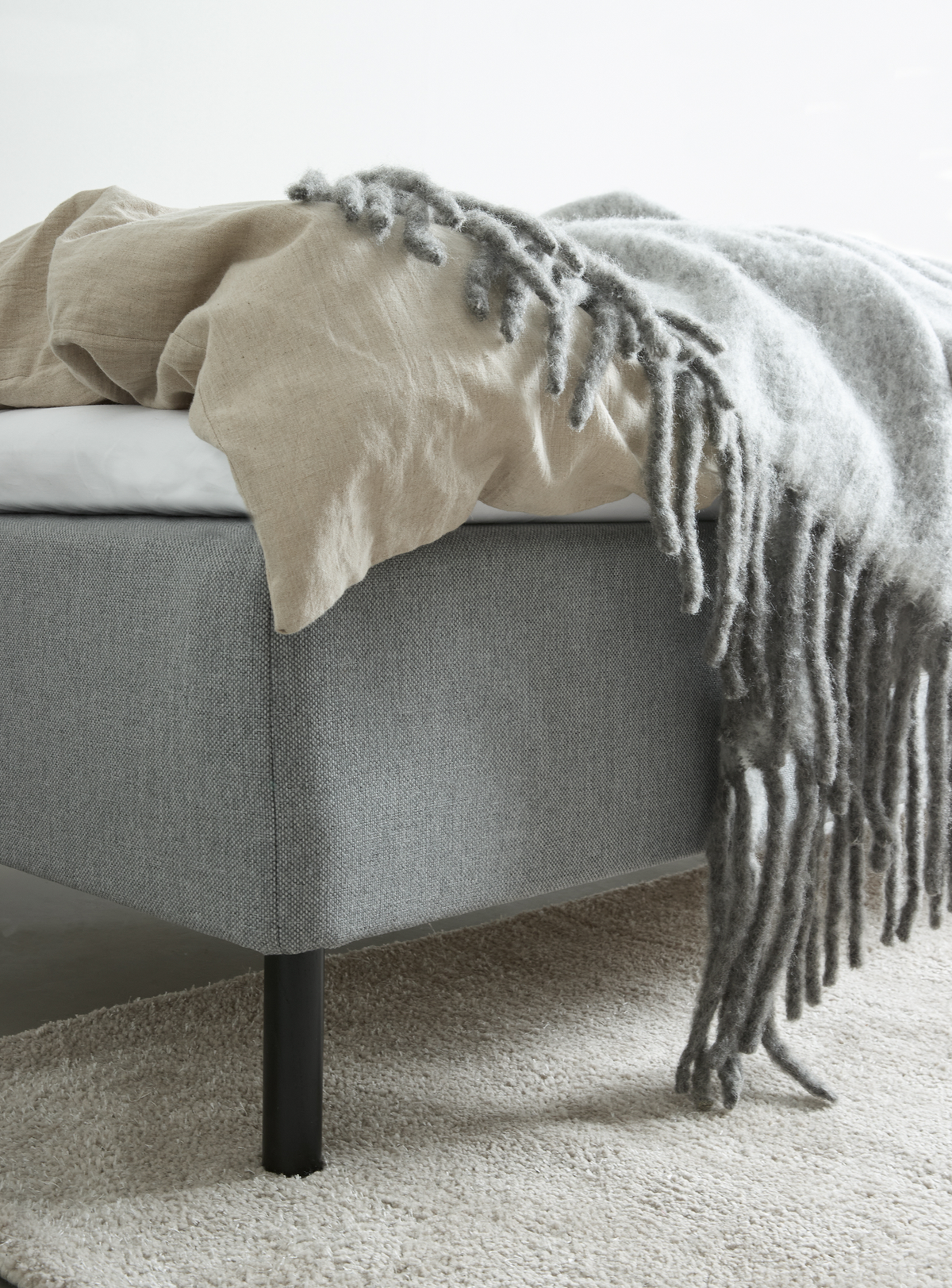 Nærbillede af sengehjørne med gråt stofbetrukket sengestel og sort ben, beige linnedyne og grå frynset plaid på lyst tæppe.