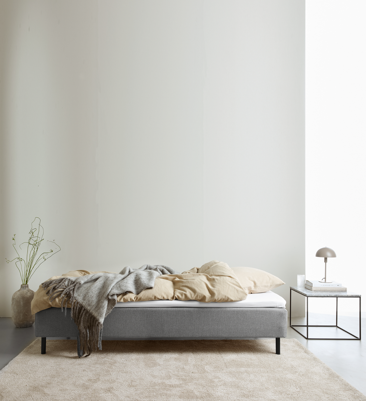Minimalistisk soveværelse med grå seng, beige sengetøj, gråt plaid, lyst tæppe og sengebord med lampe og bøger ved stort vindue.