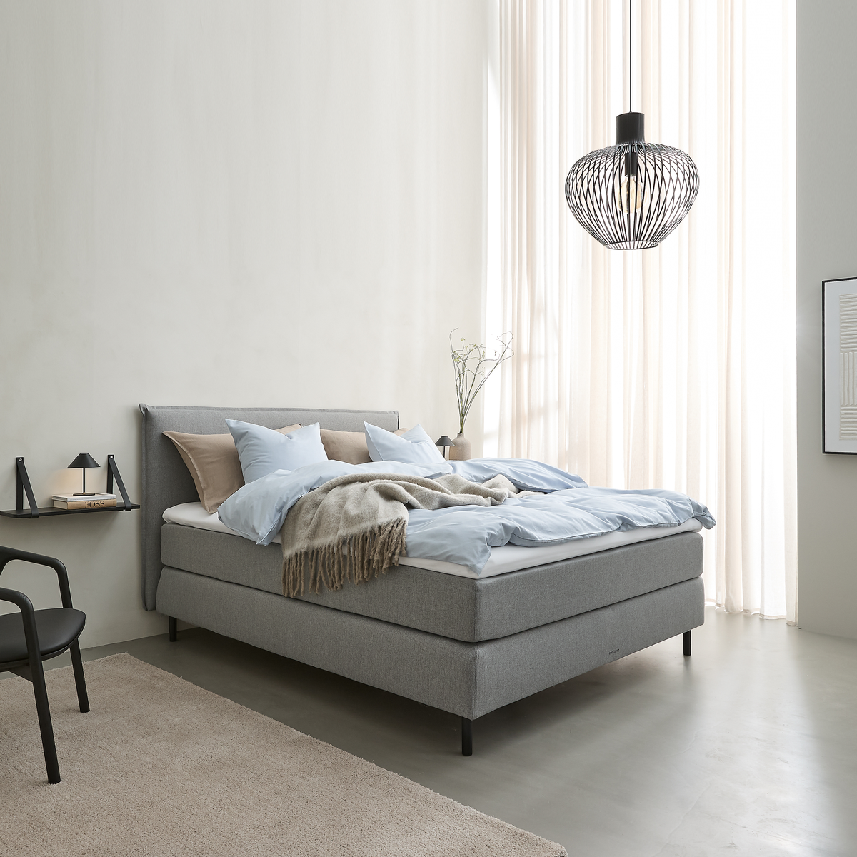 Lyst soveværelse i skandinavisk stil med grå polstret seng, lyseblåt sengetøj og beige plaid, sort trådpendel og lette gardiner, der slipper dagslys ind.