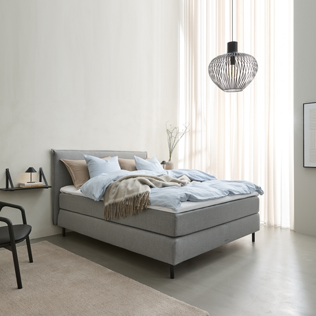 Lyst soveværelse i skandinavisk stil med grå polstret seng, lyseblåt sengetøj og beige plaid, sort trådpendel og lette gardiner, der slipper dagslys ind.