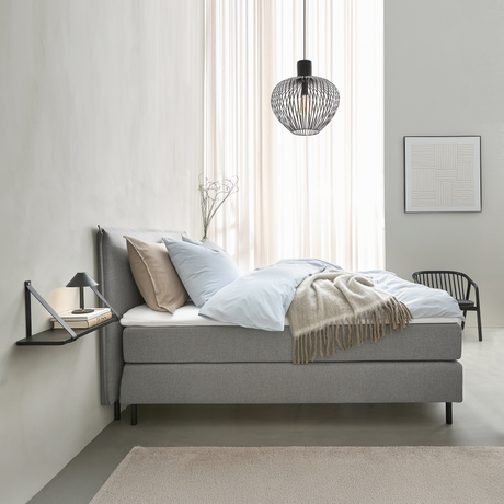 Minimalistisk soveværelse med grå seng, lyseblåt sengetøj, beige plaid, sort pendel og lille væghylde med lampe og bøger; blødt dagslys gennem lette gardiner.