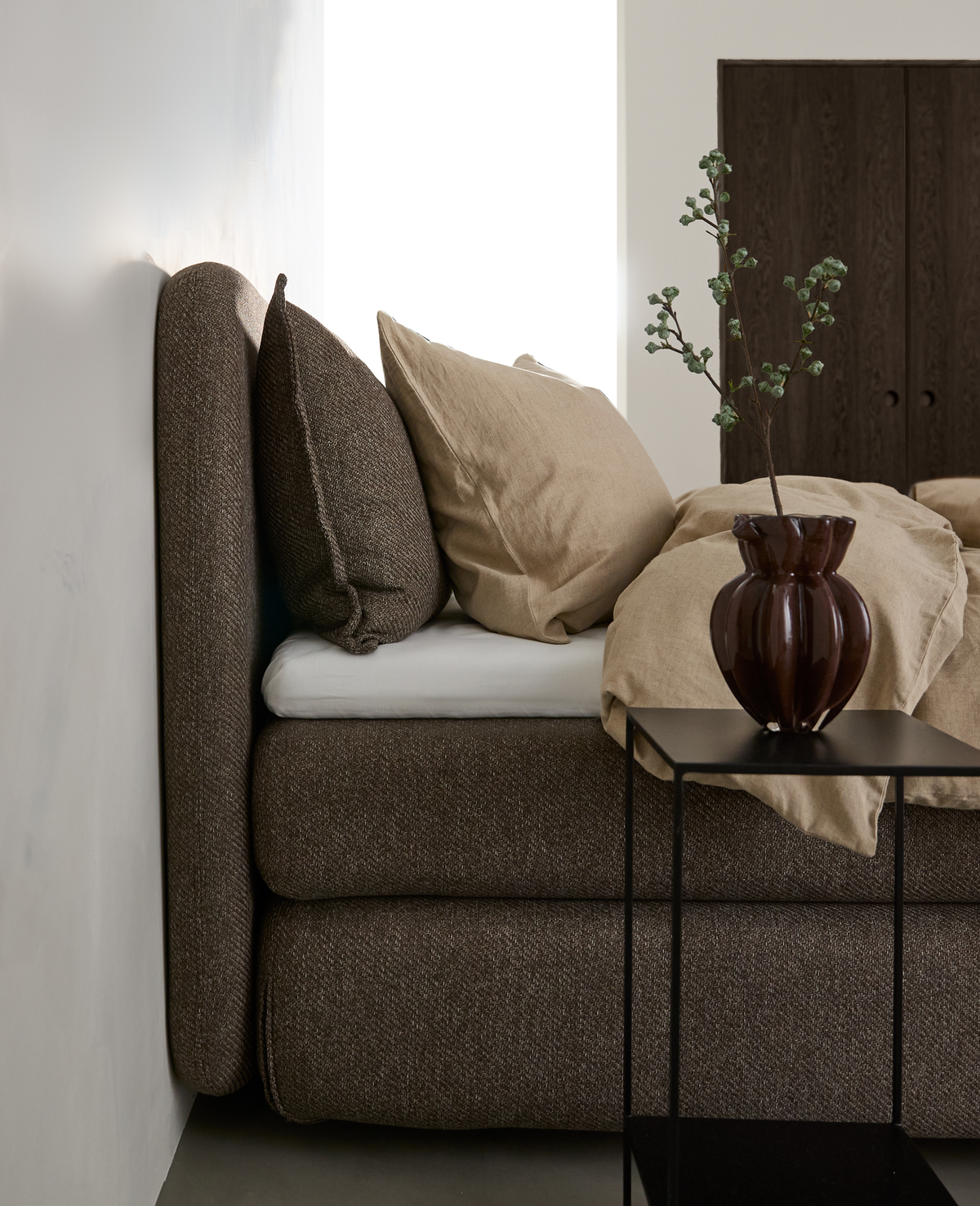 Brun polstret seng med beige sengetøj, sort sidebord med brun vase og gren i et lyst soveværelse.
