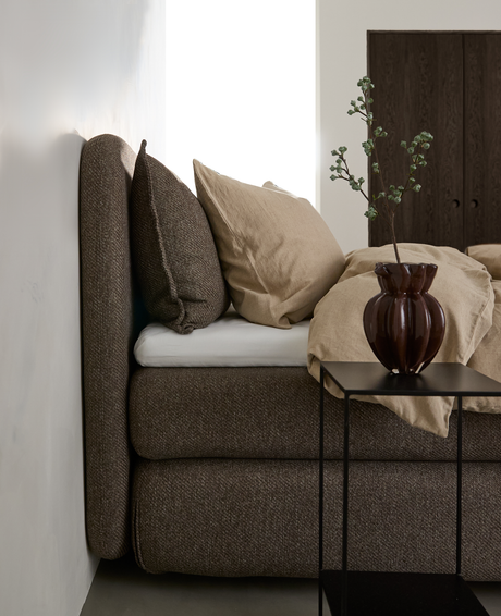 Brun polstret seng med beige sengetøj, sort sidebord med brun vase og gren i et lyst soveværelse.