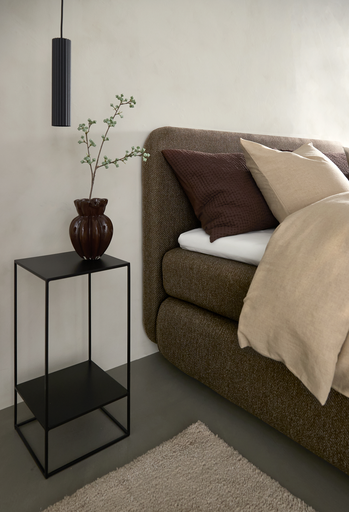 Nærbillede af soveværelse med brun polstret seng, beige og brune puder, sort metal-sidebord med brun vase og gren samt en hængende sort pendellampe.