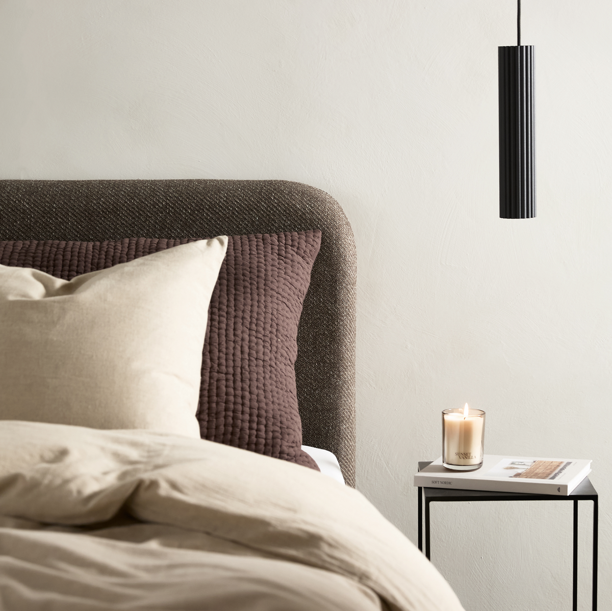 Neutralt soveværelse med beige sengetøj, brun quiltet pude, sort pendellampe og sidebord med tændt vaniljelys og bogen New Nordic.