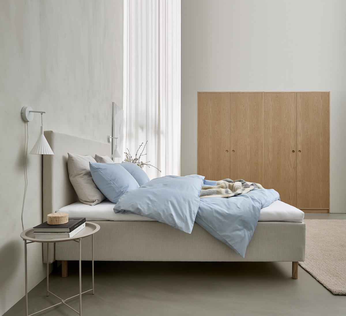 Minimalistisk soveværelse med beige polstret seng med lyseblåt sengetøj, garderobeskabe i træ, hvid væglampe og rundt natbord med bøger; blødt dagslys gennem lette gardiner.