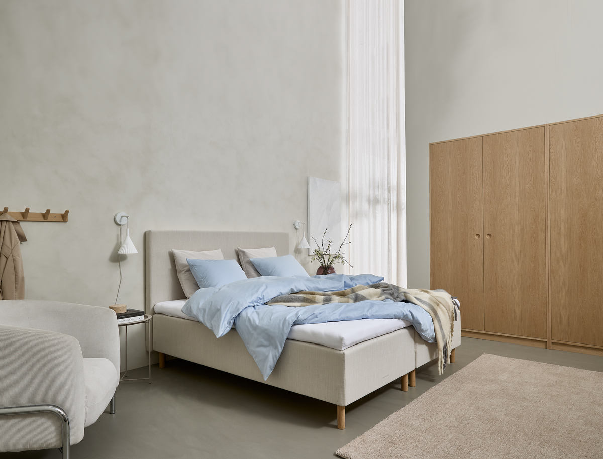 Lyst, minimalistisk soveværelse med beige polstret seng, lyseblåt sengetøj, væglamper, træskab og blødt tæppe ved et højt vindue med lette gardiner.