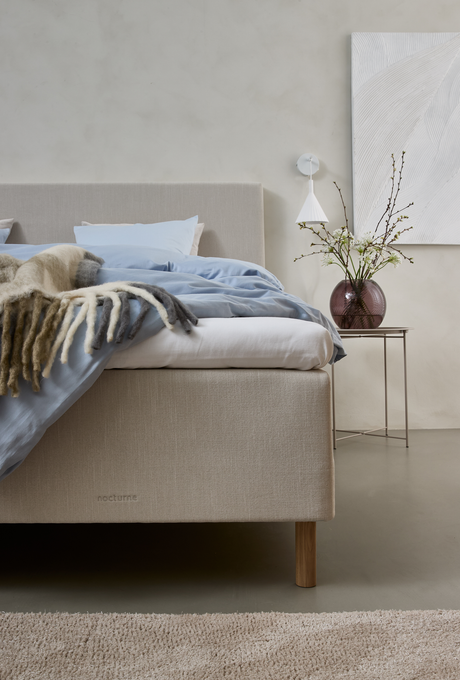 Beige polstret seng med blåt sengetøj og uldtæppe, nocturne på sengebunden; sidebord med rosa glasvase med grene, væglampe og hvidt tekstureret kunst i et roligt, neutralt soveværelse.