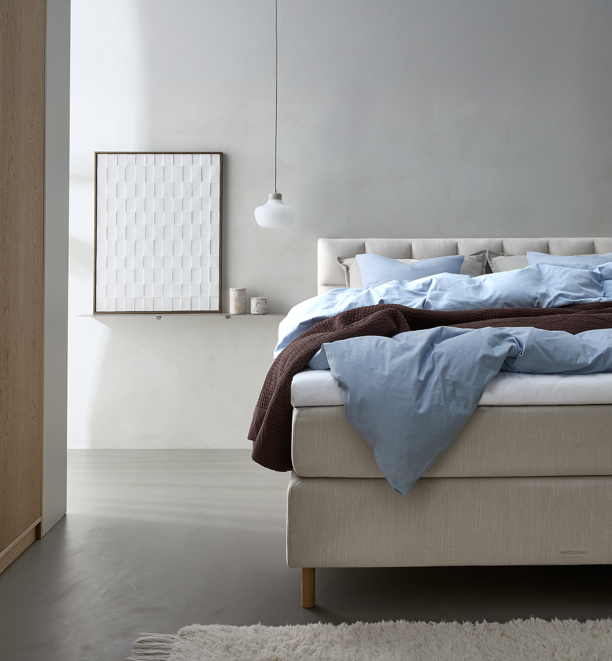 Lyst soveværelse med beige kontinentalseng redt med lyseblåt sengetøj og brun plaid, hvid pendel og billede på hylde.