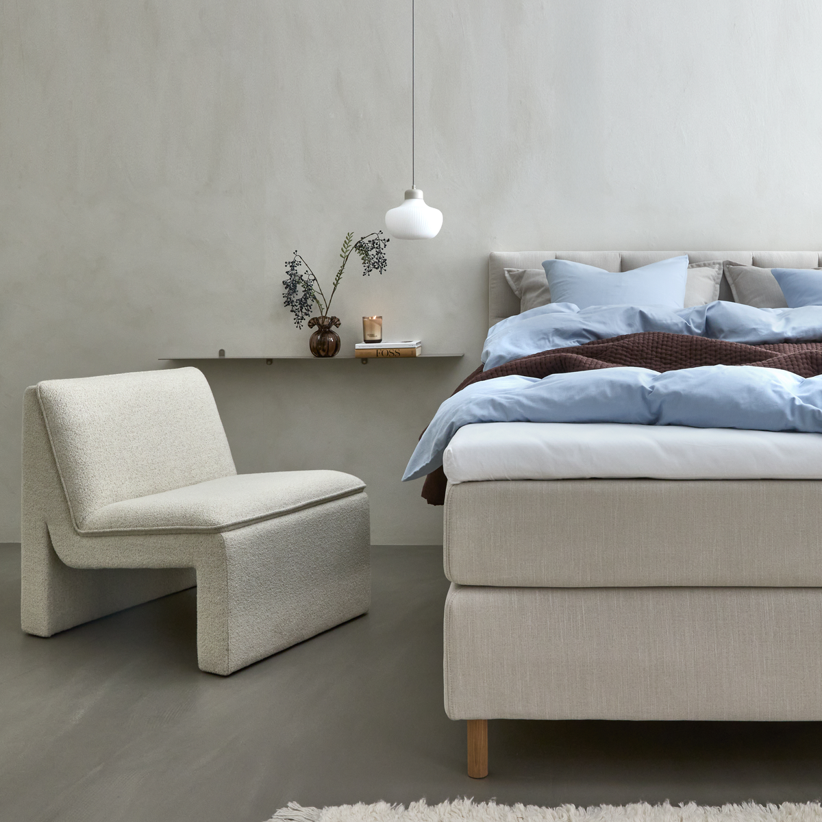 Rolig skandinavisk soveværelsesscene med beige seng, lyseblåt sengetøj og brun plaid, skulpturel lys lænestol, smal hylde med vase, bog og lys samt hvid pendellampe.