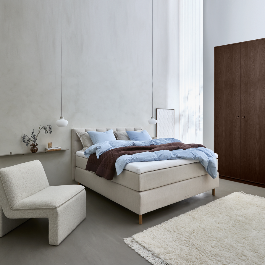 Lyst skandinavisk soveværelse med beige polstret seng, lyseblåt sengetøj og brun plaid, to pendler, lav lounge-stol, vase med grene på hylde, lette gardiner, hvid langhåret tæppe og mørkt klædeskab.