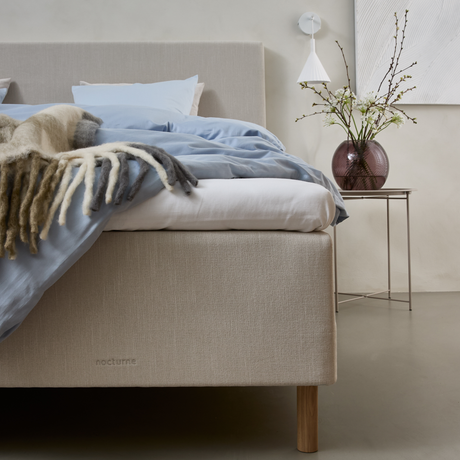 Beige seng med blåt sengetøj, uldplaid og diskret mærkning nocturne; glasvase med grene på et sidebord og hvid væglampe i et lyst soveværelse.