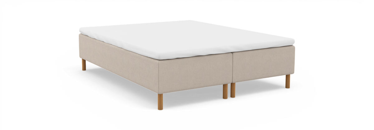 Beige kontinentalsengbund med hvid topmadras og runde træben, frilagt på hvid baggrund.