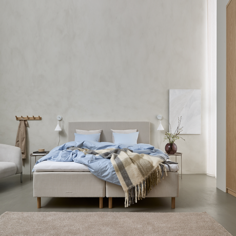 Lyst soveværelse med beige kontinentalseng, lyseblåt sengetøj og ternet plaid over sengen, væglamper og vase med grene på sengebordet.