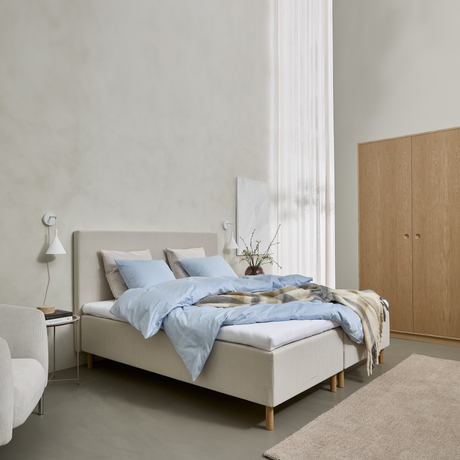Lyst soveværelse med beige seng, lyseblåt sengetøj og plaid ved fodenden, væglamper, lette gardiner og et skab i træ.