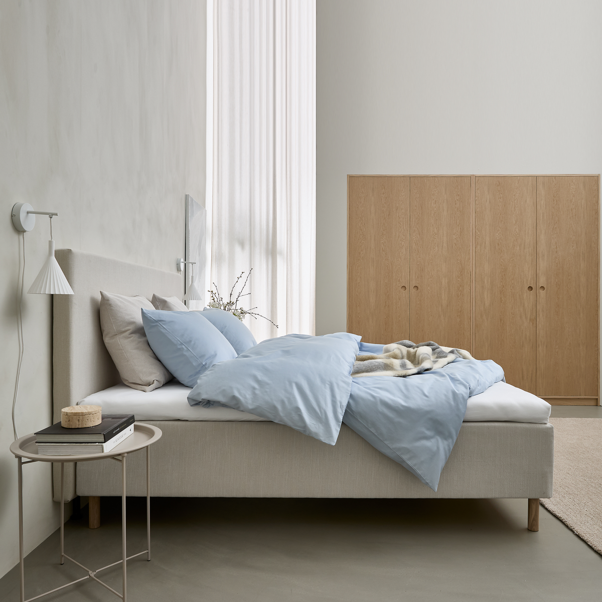 Lyst, minimalistisk soveværelse med beige seng, lyseblåt sengetøj og puder, væglampe, lille sidebord med bøger og garderobeskabe i lys eg.