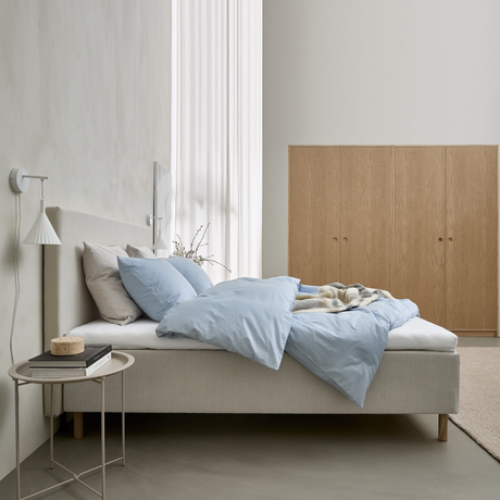 Lyst, minimalistisk soveværelse med beige seng, lyseblåt sengetøj og puder, væglampe, lille sidebord med bøger og garderobeskabe i lys eg.